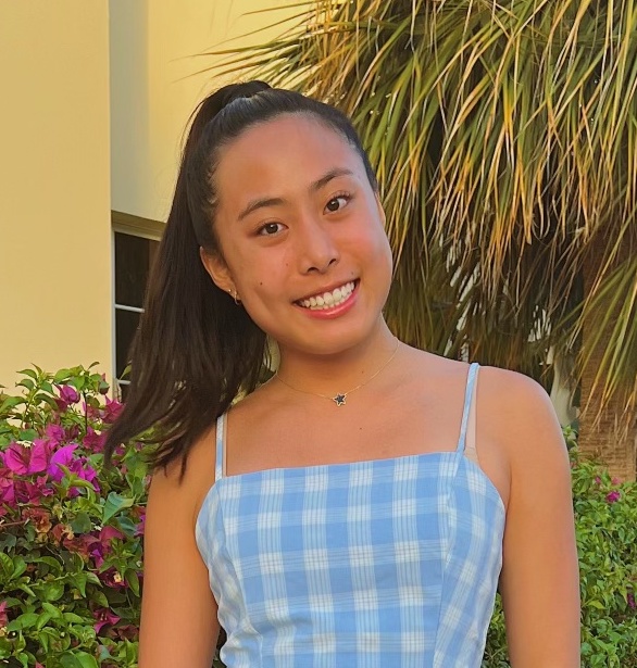 A picture of Megan smiling and looking at the audience. Her black hair is in a ponytail, and she is wearing a blue plaid dress. She is stading in front of a palm tree.