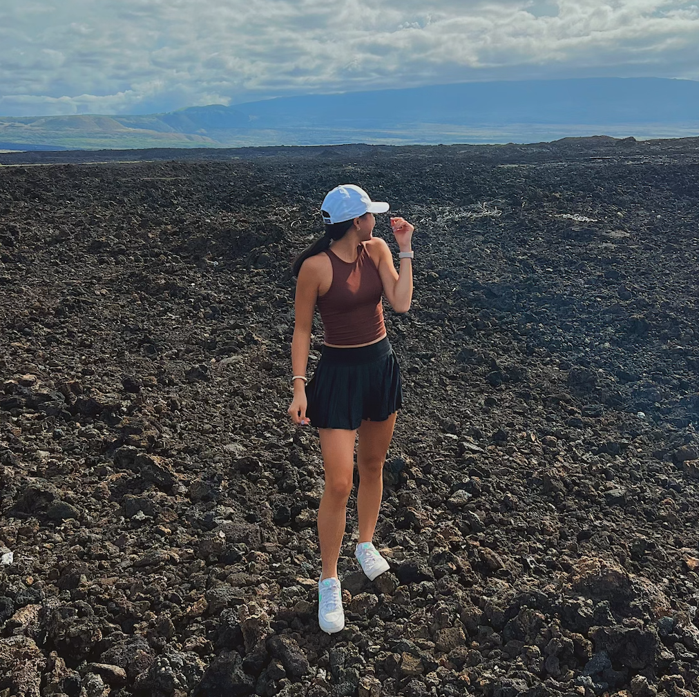 A picture of Megan standing on lava rocks. She is wearing a brown tank top, black skirt, and white baseball cap. Her head is turned away from the camera.